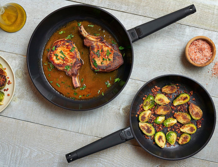 Zavor noir 10 & 12 inch skillet set with black handles with food on the skillets and around them on a light colored wooden surface