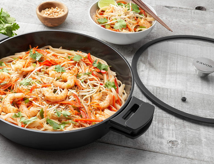 Stir fry inside a Zavor Noir pan next to a Zavor lid and bowl of stir fry displayed on a wooden surface