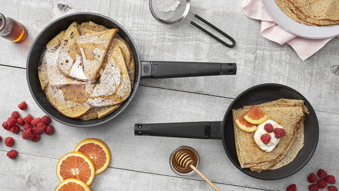 Top view of set of Zavor noir 8 and 10 inch skillet with French toast and crepes next to baking materials and fruit displayed on a wooden surface