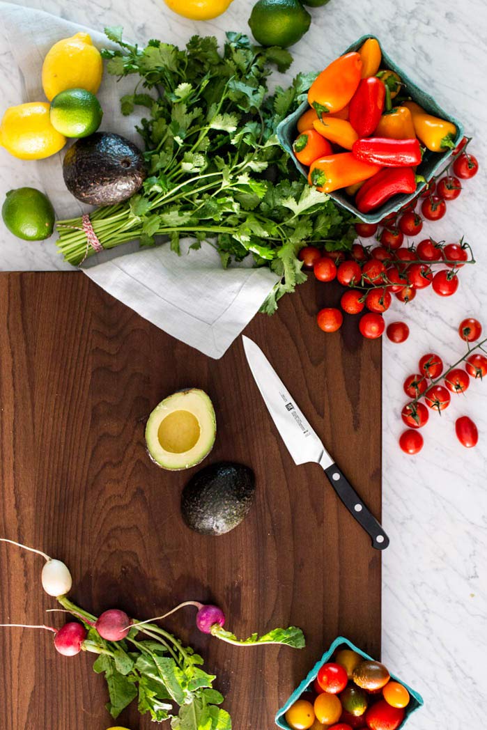Zwilling 5.5 inche pro ultimate prep tomato utility knife on a wooden cutting board next to lemons, avocados, limes, peppers, tomatoes, and radishes on a marble countertop