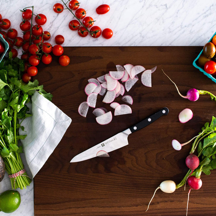 Zwilling pro ultimate 5.5 inch prep tomato utility knife on a wooden cutting board next to tomatoes, herbs, and radish